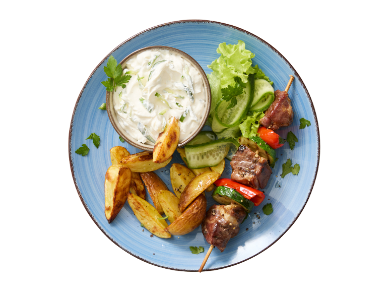 Ražnjić od mesa i povrća, pečeni krumpir i tzatziki na plavom tanjuru.