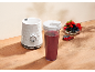Bijeli blender s crvenim voćnim smoothiejem i zdjelom bobičastog voća na drvenom stolu.