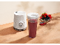 Bijeli blender s crvenim voćnim smoothiejem i zdjelom bobičastog voća na drvenom stolu.