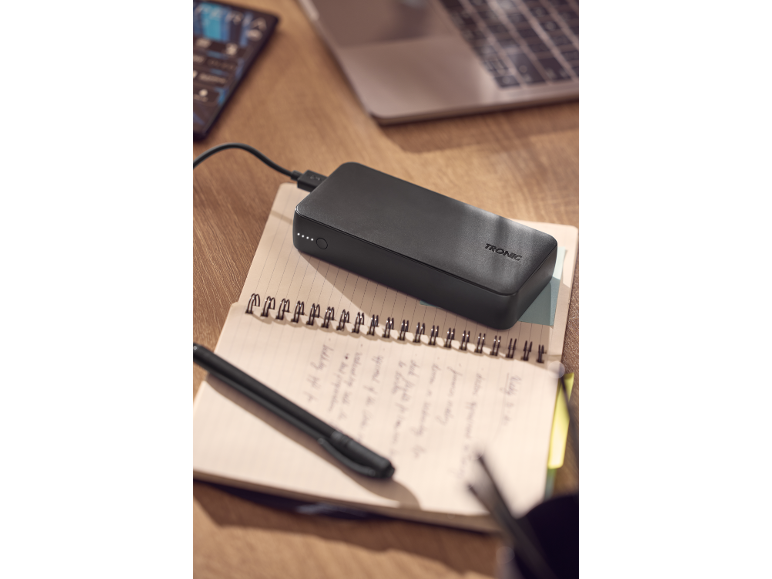 Crni power bank puni se na otvorenoj bilježnici s olovkom, laptop u pozadini.