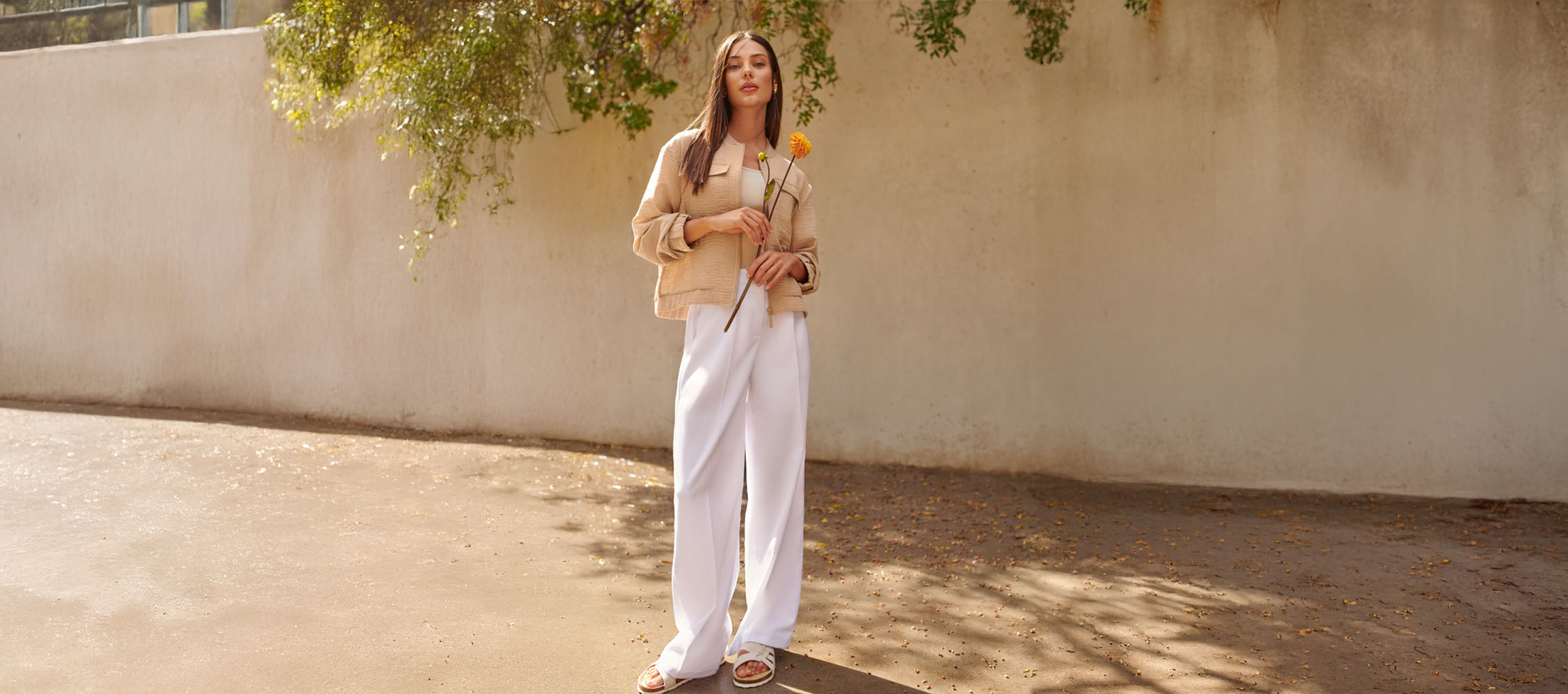 Žena u bež jakni, bijelim hlačama i sandalama, drži žuti cvijet.