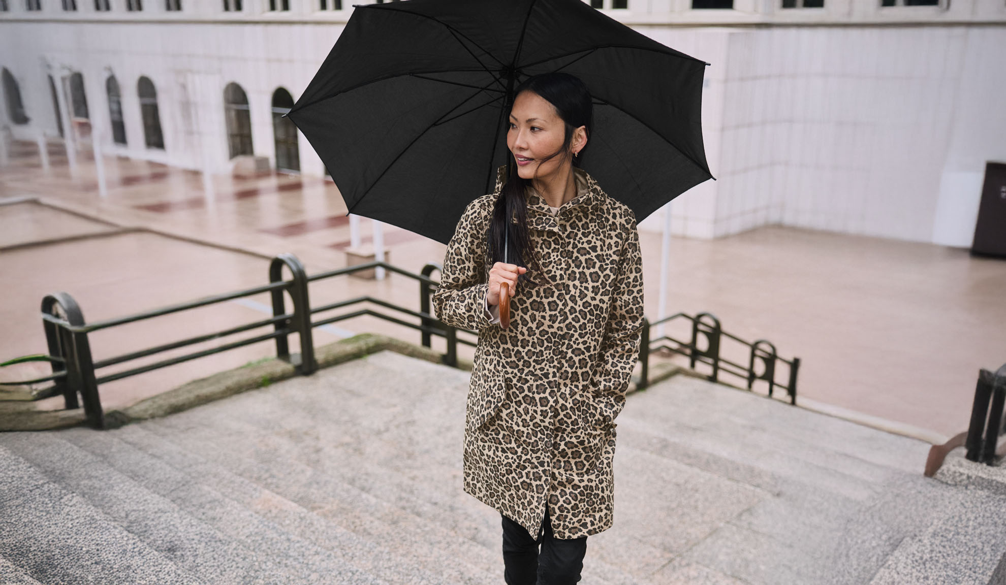 Žena u kaputu s leopard uzorkom drži crni kišobran na stubištu.