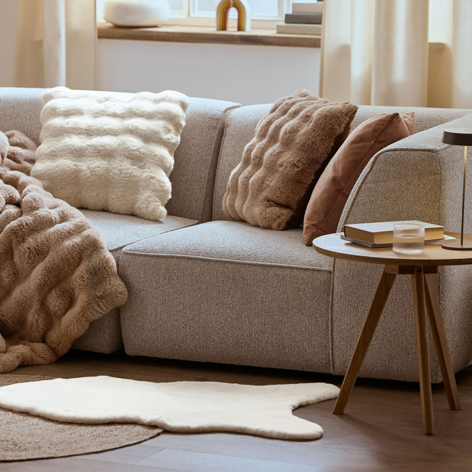 Siva modularna sofa s pahuljastim jastucima i dekom, pored drvenog bočnog stolića.