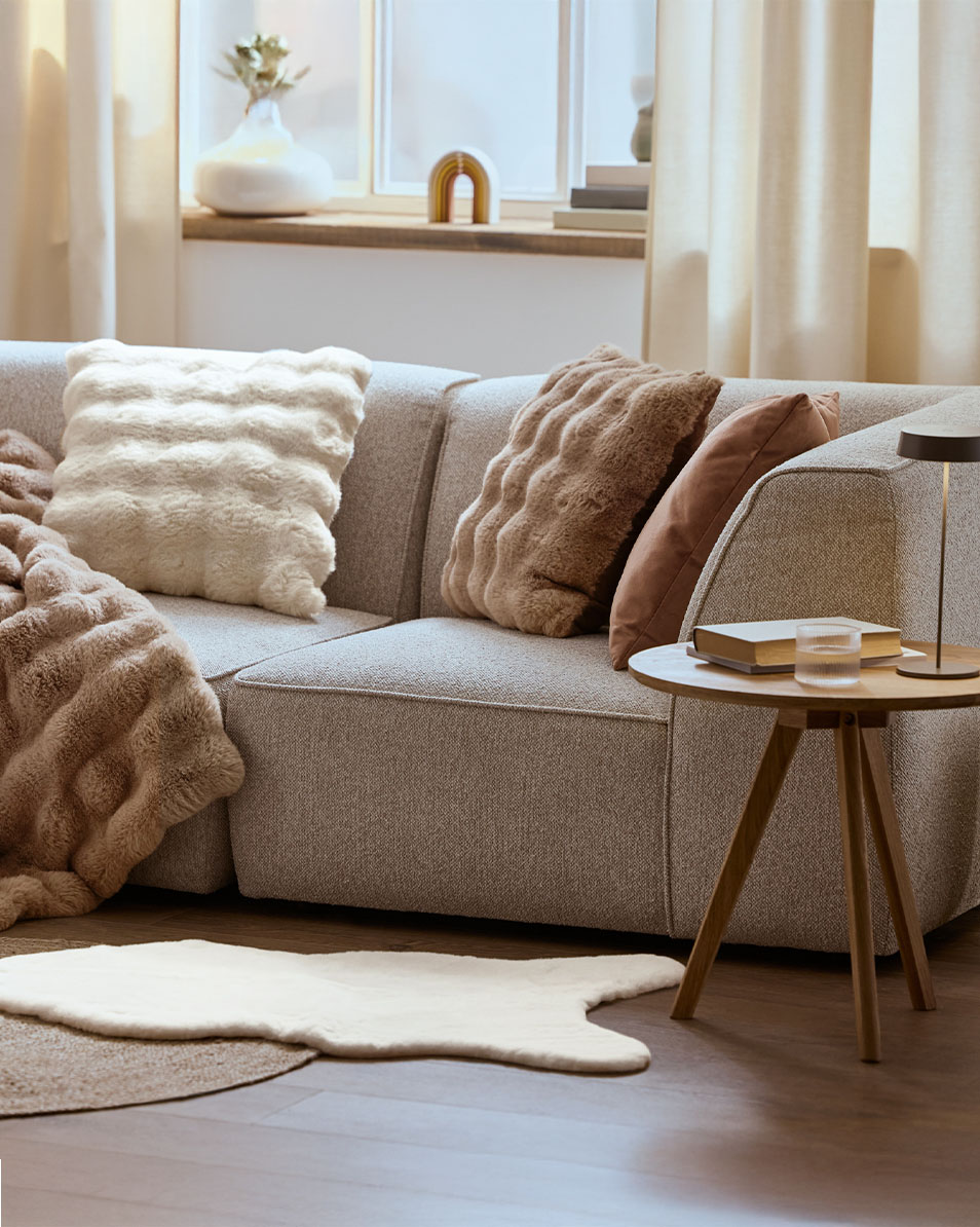 Siva modularna sofa s pahuljastim jastucima i dekama, drveni bočni stolić.