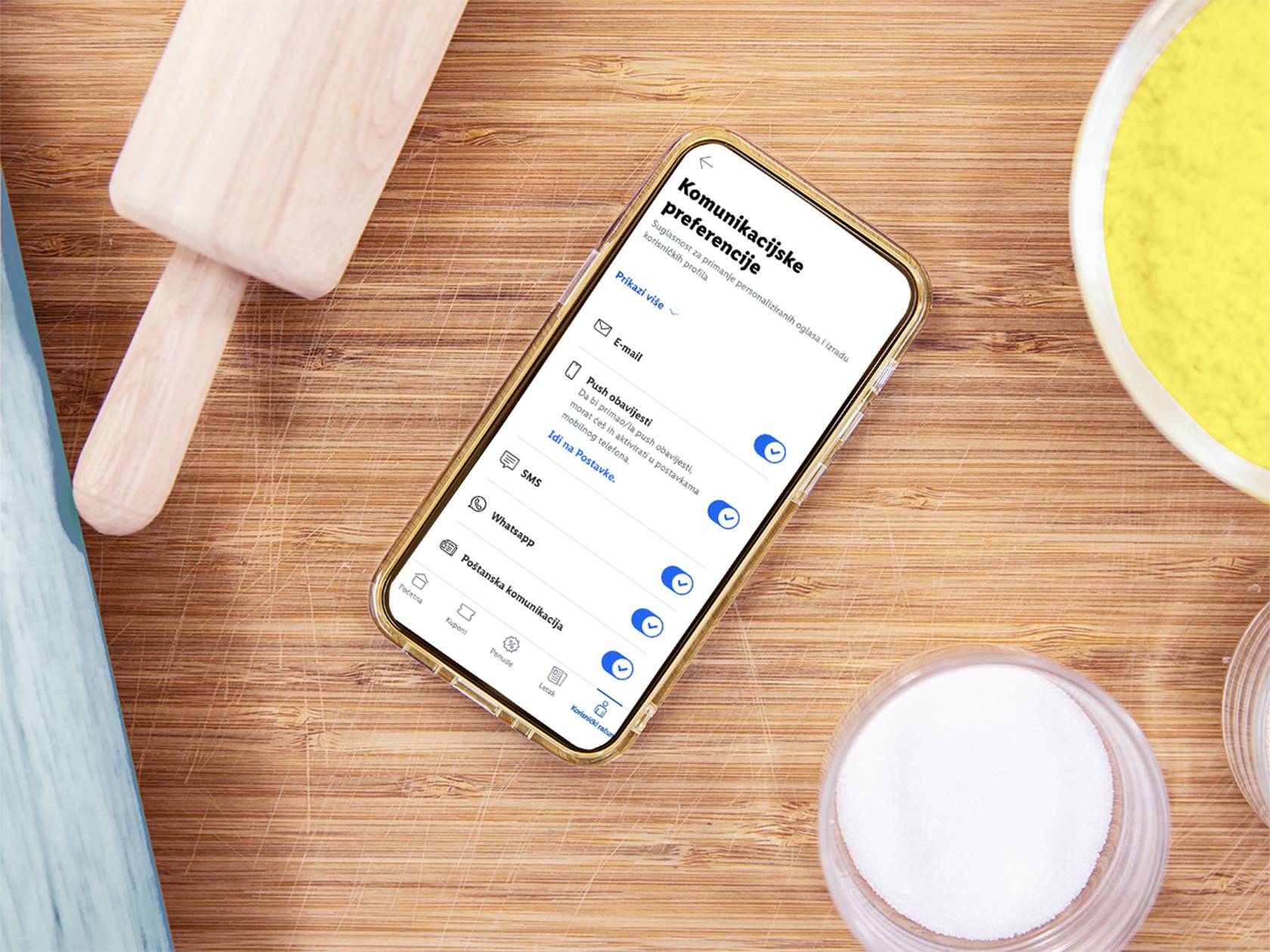 Pametni telefon s aplikacijom Lidl Plus na drvenoj dasci za rezanje, okružen kuhinjskim priborom.