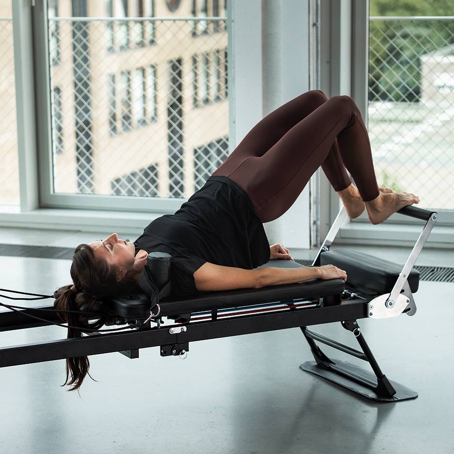 Žena vježba Pilates na reformer spravi, noge podignute.