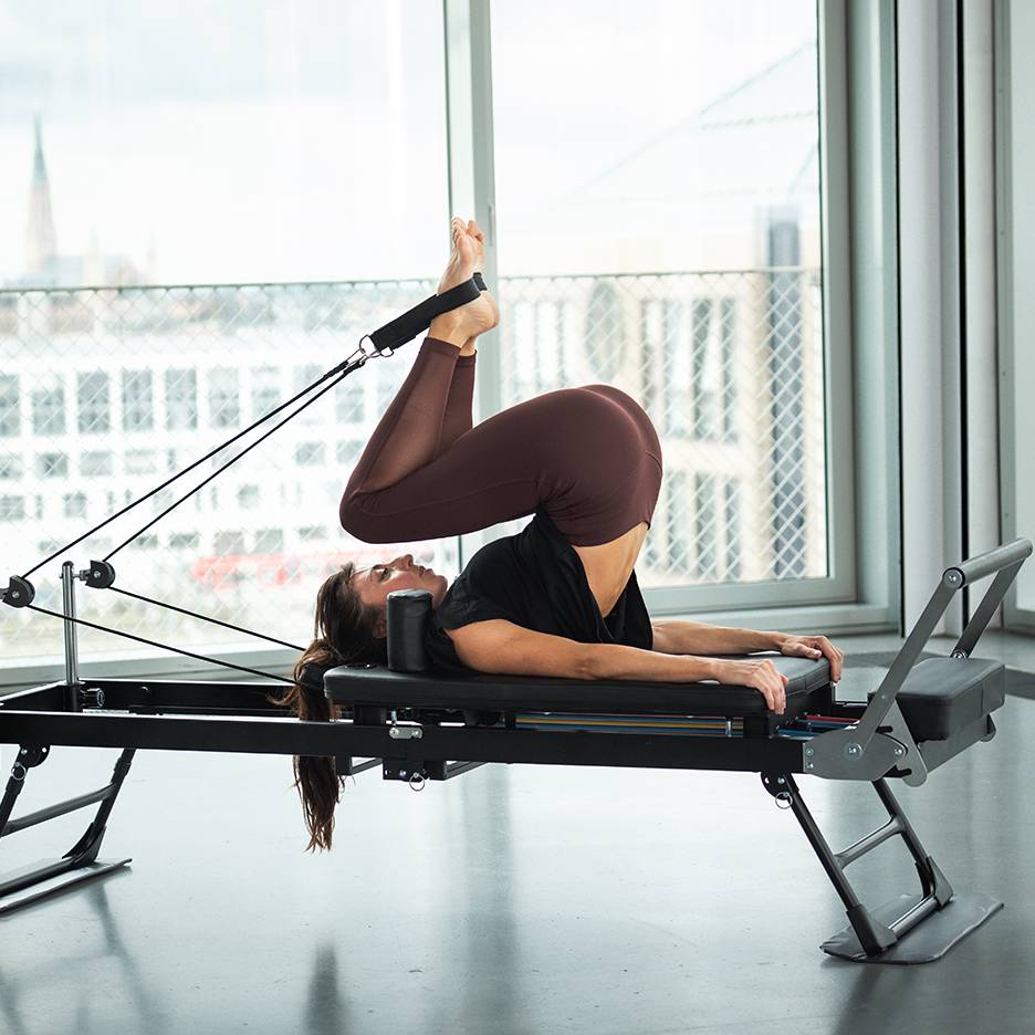 Žena vježba pilates na reformer spravi s podignutim nogama i stopalima u trakama.