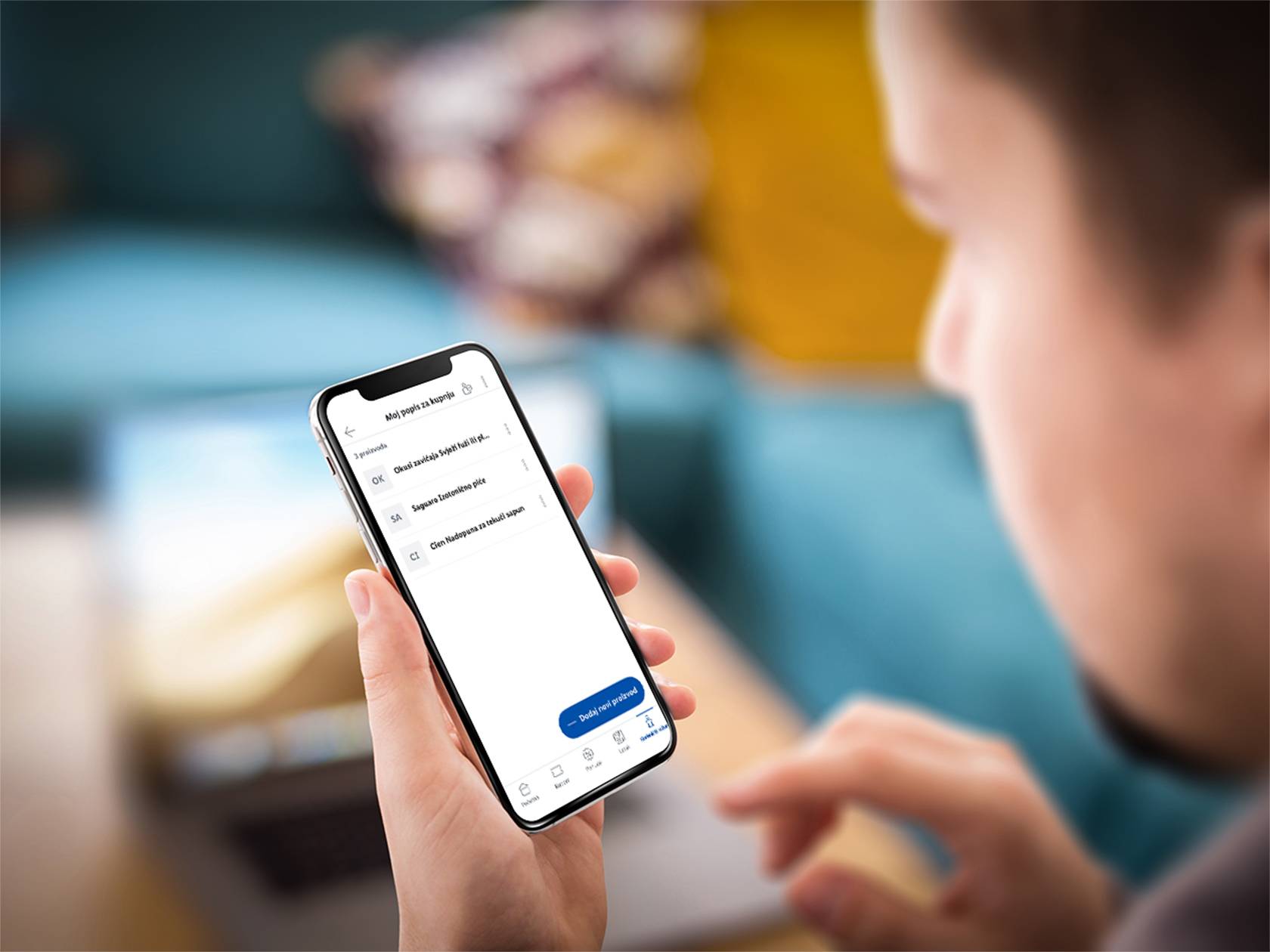 Osoba drži pametni telefon s aplikacijom za kupovinu koja prikazuje popis proizvoda poput 'Okusi zavičaja Svježi fuži', 'Saguaro Izotonično piće' i 'Cien Nadopuna za tekući sapun'.