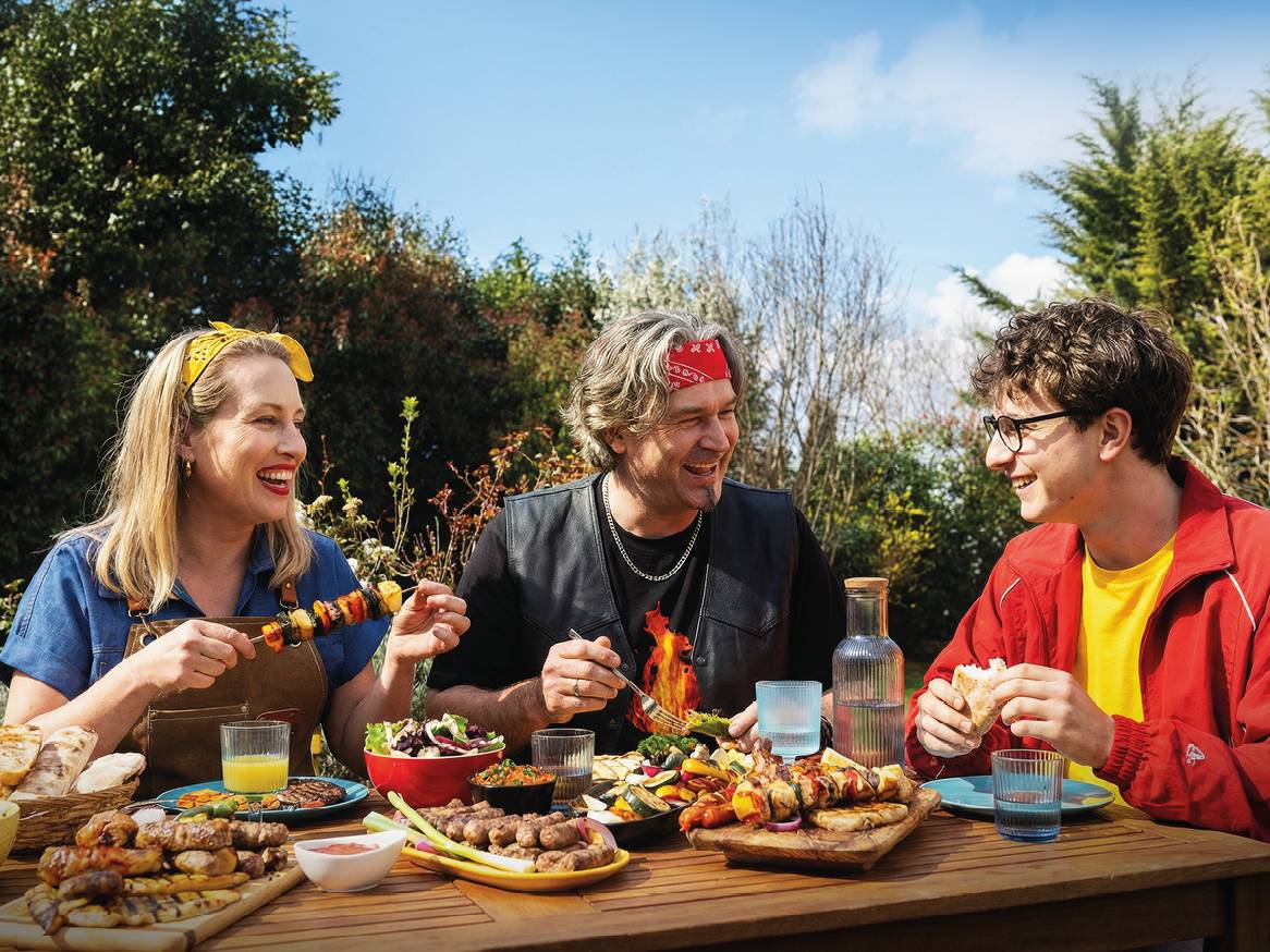 Obitelj uživa u roštilju na otvorenom s mesom, povrćem i salatama na drvenom stolu.