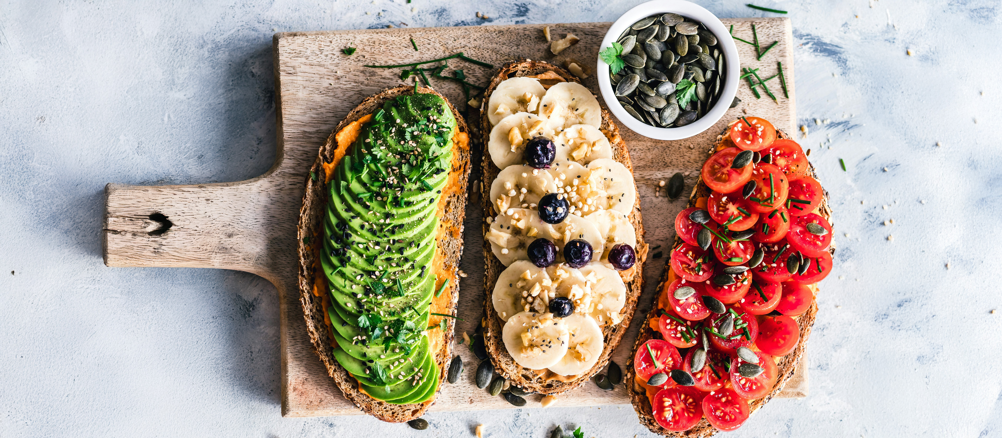 Tri tosta s avokadom, bananom i borovnicama te rajčicama na drvenoj dasci.