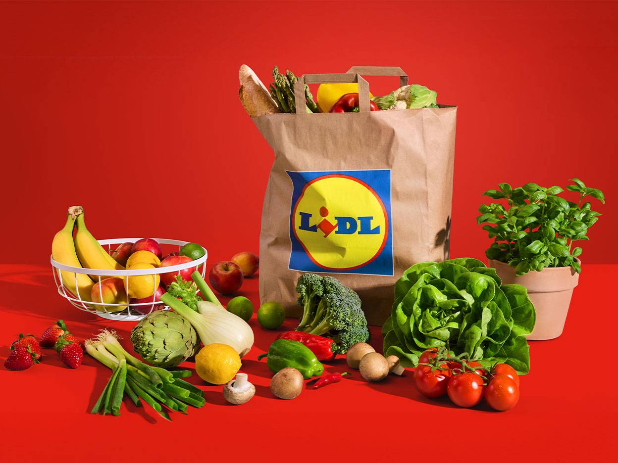 Raznovrsno svježe voće i povrće, uključujući banane, jabuke, zelenu salatu i rajčice, s Lidl vrećicom.
