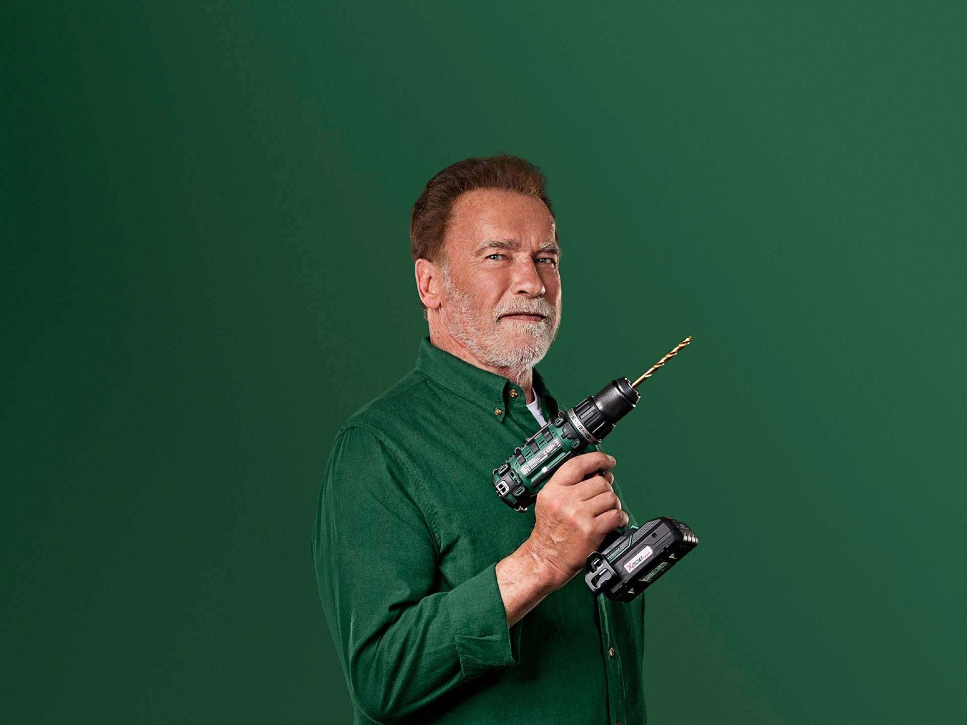 Arnold Schwarzenegger drži zelenu električnu bušilicu na zelenoj pozadini.