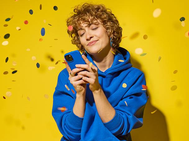 Žena u plavoj majici s kapuljačom drži pametni telefon dok pada konfeti.