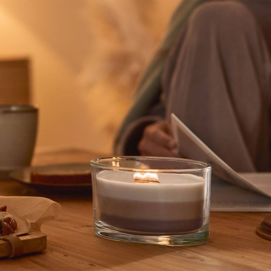 Upaljena troslojna svijeća u čaši, s osobom koja čita u pozadini.