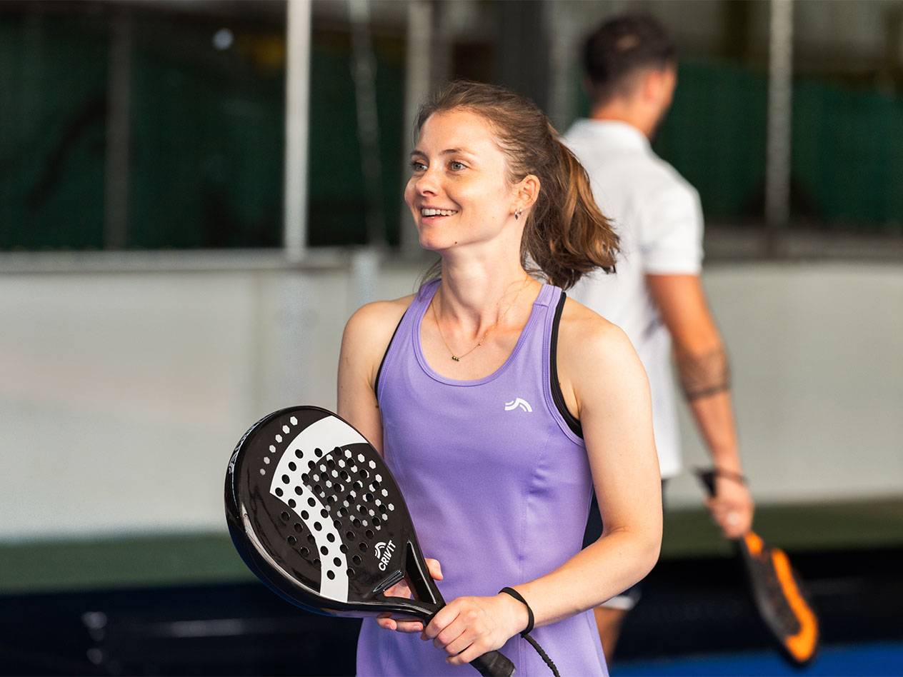 Žena u ljubičastoj sportskoj haljini drži reket za padel, muškarac u pozadini.