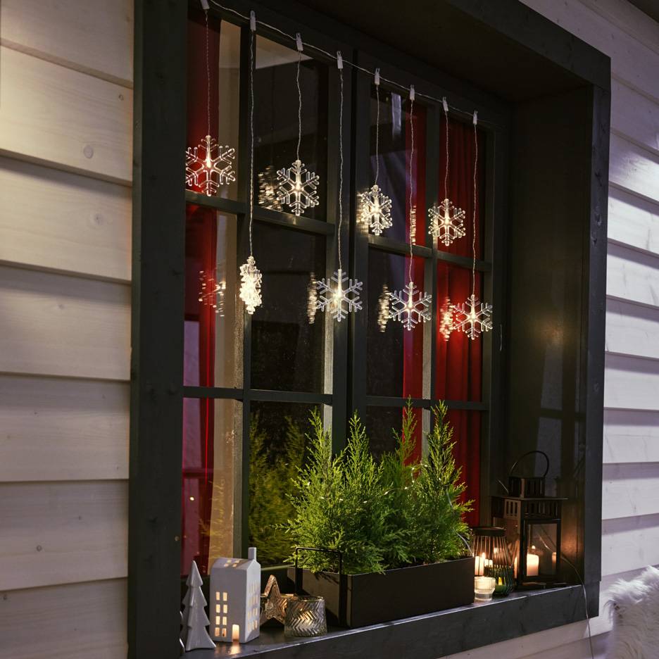 Svjetleće pahuljice vise u prozoru s malim jelkama i svijećama na prozorskoj dasci.