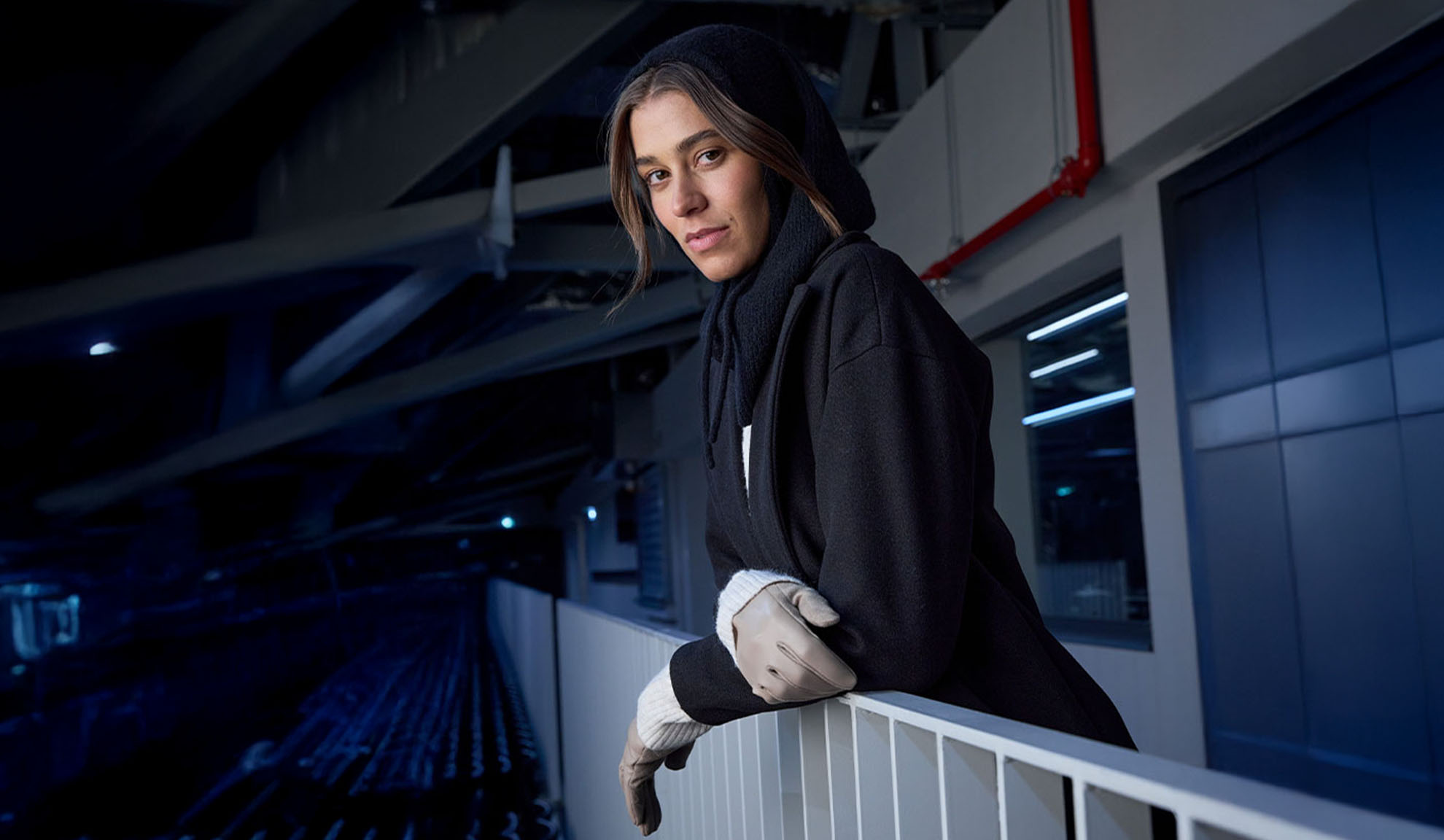 Žena u crnom kaputu i marami s kapuljačom, naslonjena na ogradu u modernom okruženju.