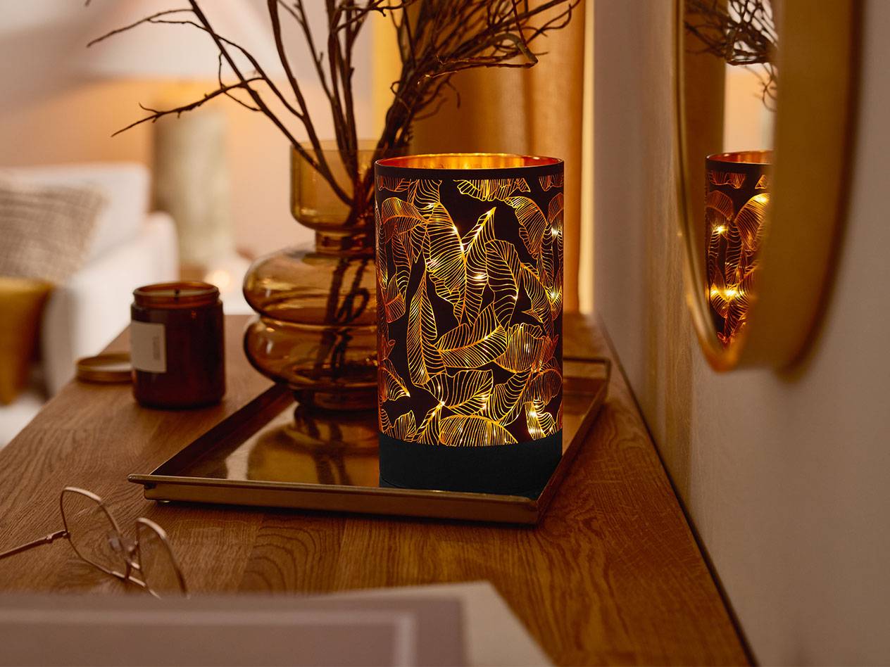 Dekorativna LED svjetiljka s uzorkom lišća, vaza s granama i svijeća na drvenom stolu.