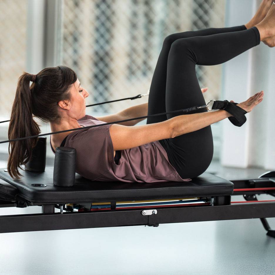 Žena vježba pilates na reformer spravi, u crnoj sportskoj odjeći.
