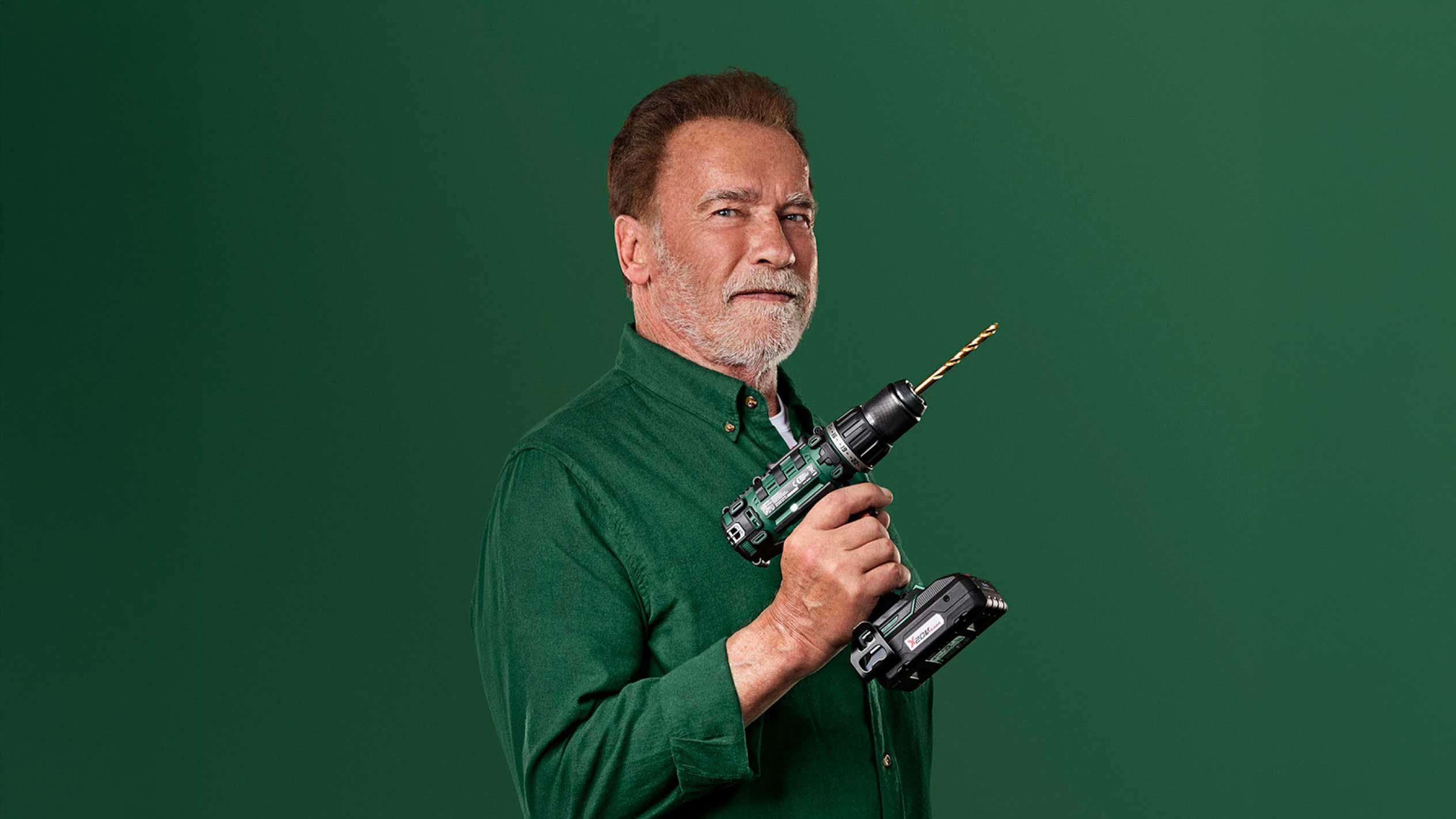 Arnold Schwarzenegger drži zelenu bežičnu bušilicu sa zlatnim svrdlom.