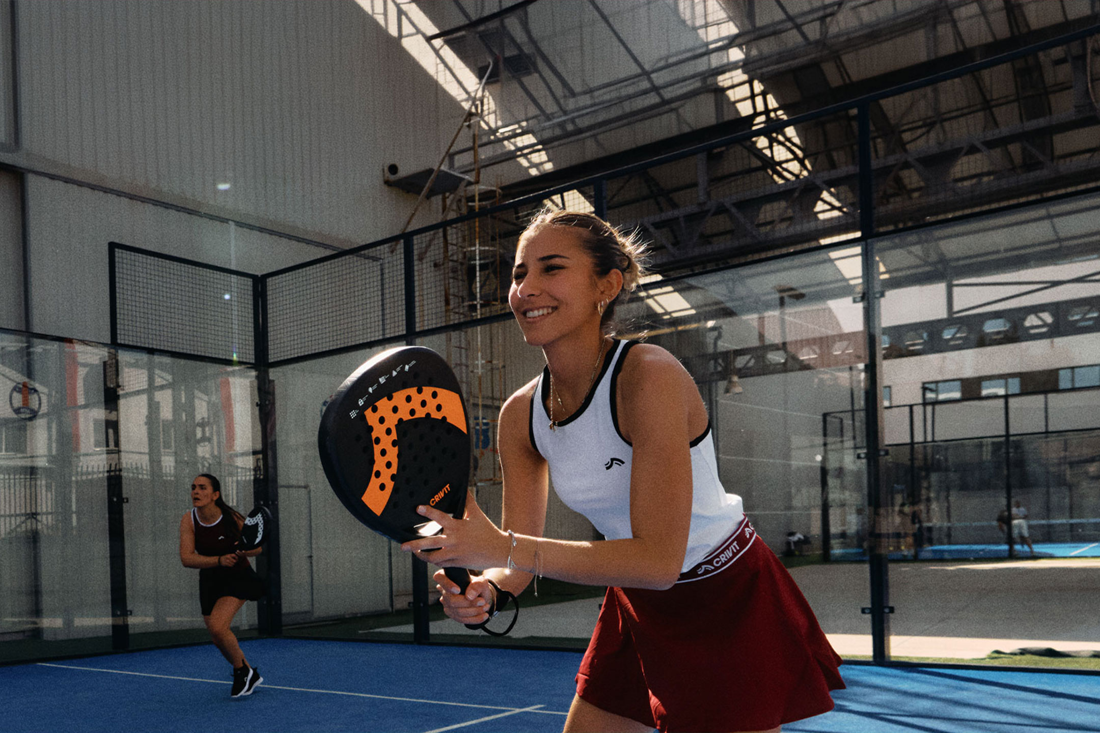 undefined: Dvije žene igraju padel tenis, jedna se smiješi i drži reket, na plavom terenu.