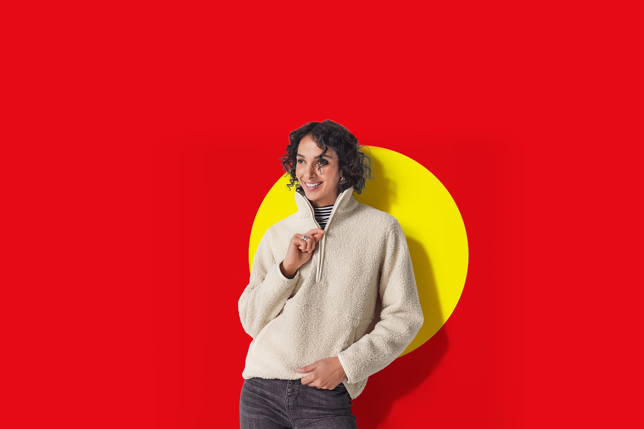 Žena u bež flis puloveru i tamnim trapericama, nasmijana, na crvenoj pozadini sa žutim krugom.