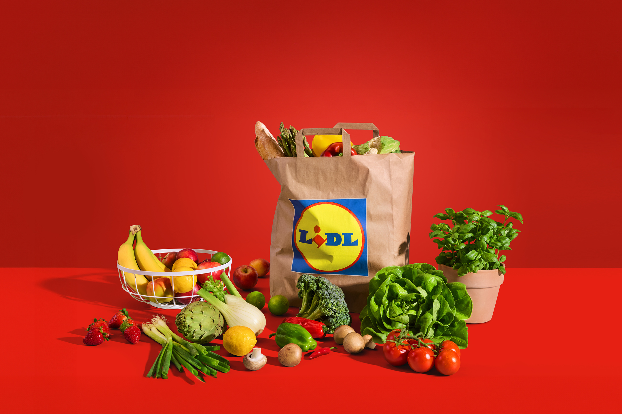 Papirnata vrećica s namirnicama i svježim voćem i povrćem na crvenoj pozadini.