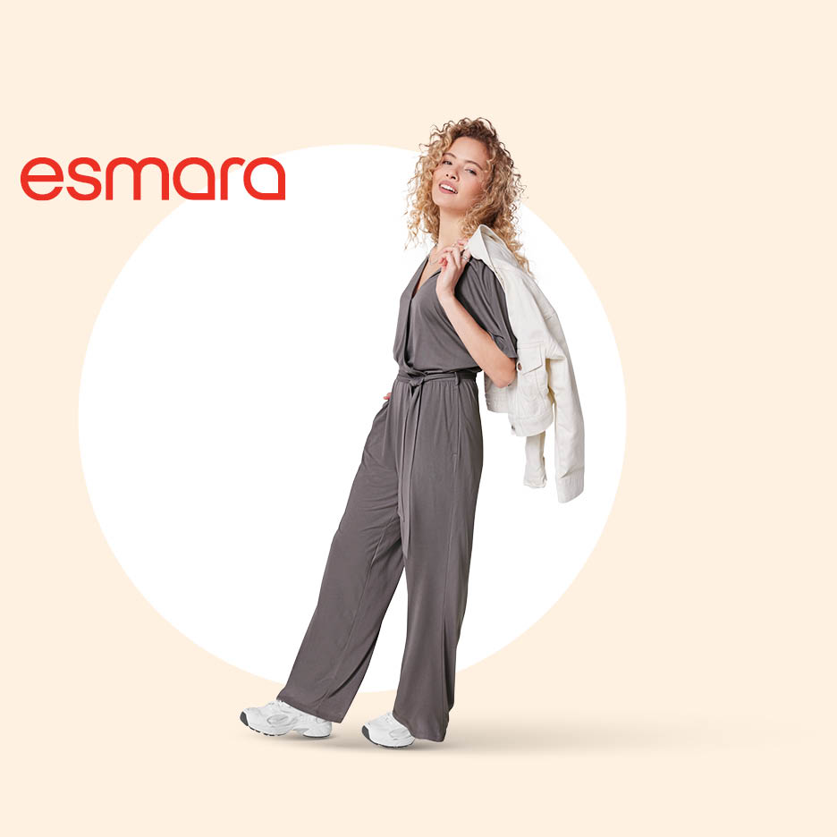 Žena u tamnosivoj kombinezonu s bijelom jaknom prebačenom preko ramena, nasmijana.