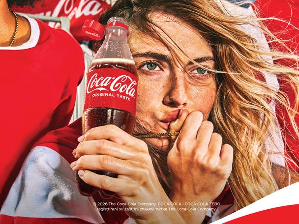 Mlada žena drži bocu gaziranog pića, dok zviždi, u pozadini je muškarac u crvenoj majici.