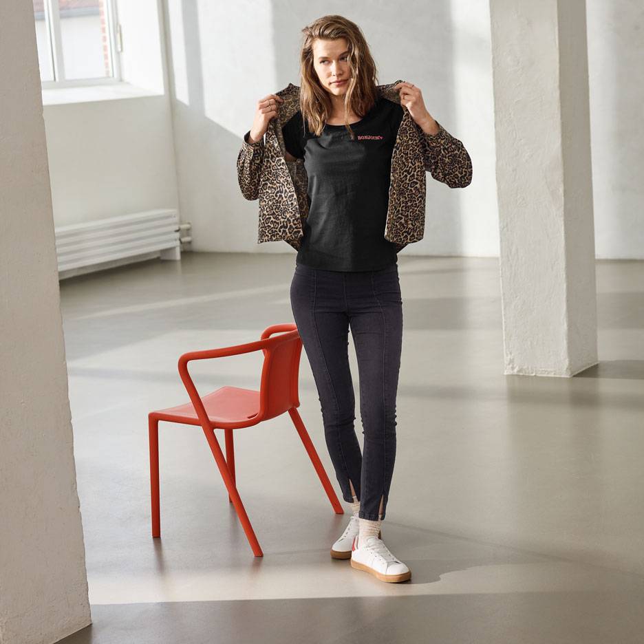 Žena u crnoj majici s natpisom, trapericama i jakni s leopard uzorkom, stoji pored crvene stolice.