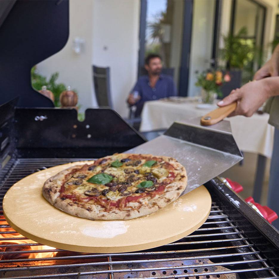 Pizza na kamenu za pizzu na roštilju, s lopatom za pizzu i muškarcem u pozadini.