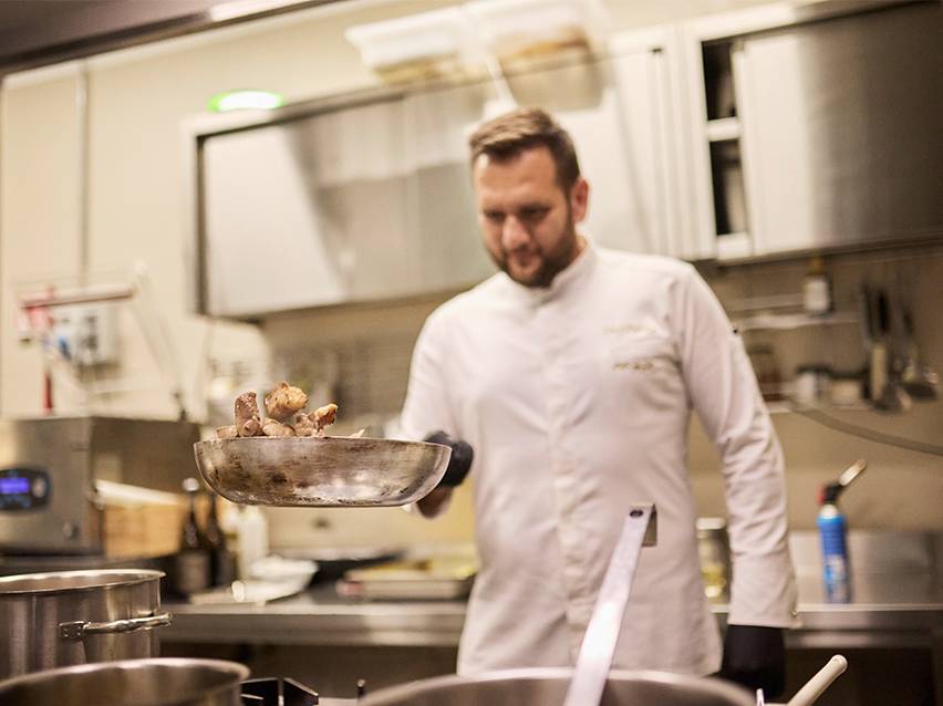 Kuhar u bijeloj kuharskoj jakni baca hranu u tavi.