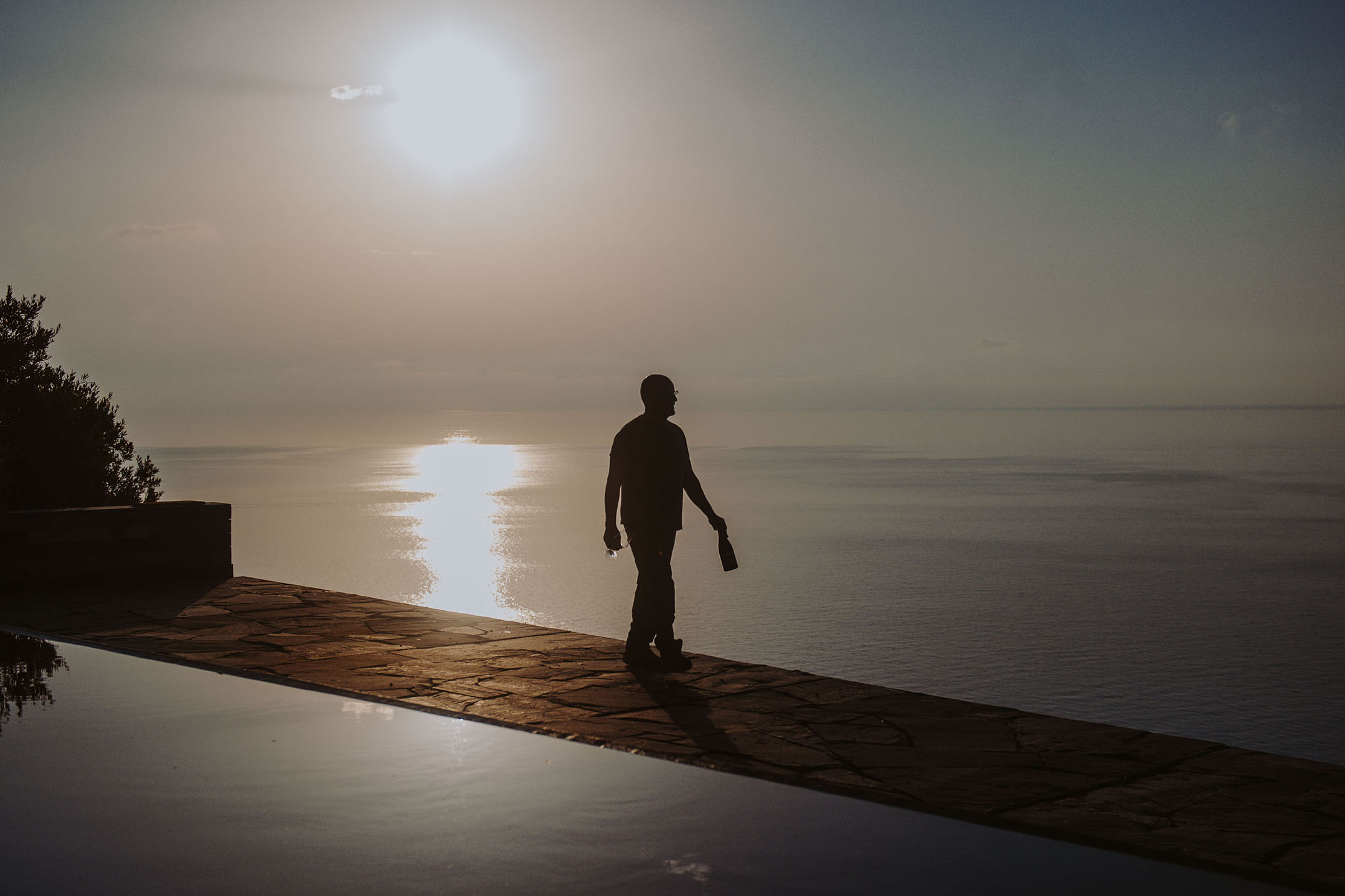 Silueta osobe s bocom i čašom pored beskonačnog bazena pri zalasku sunca