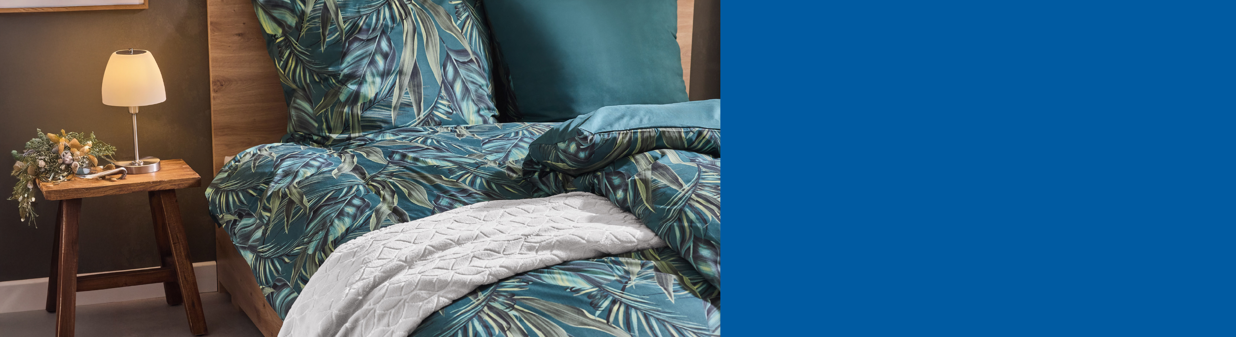 Posteljina s uzorkom tropskog lišća i jednobojni jastuci na krevetu, pored kojeg je drveni stolić s lampom.