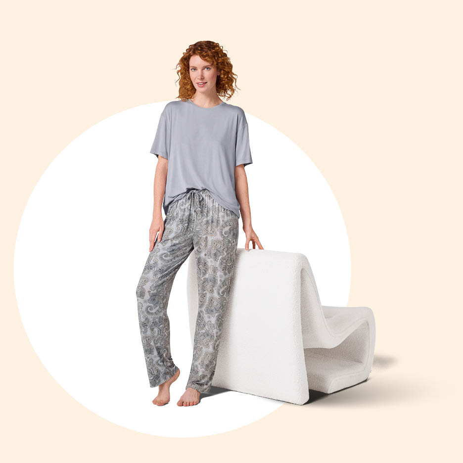 Žena u sivoj majici i sivim hlačama s uzorkom, naslonjena na bijelu stolicu.