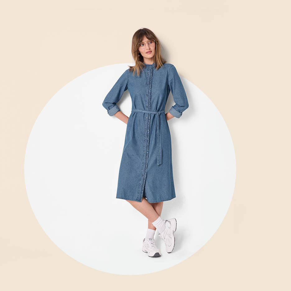 Žena u traper haljini s remenom, bijelim čarapama i tenisicama, stoji u krugu.