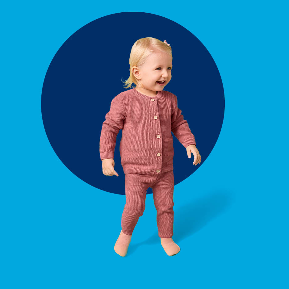 Sretna beba u ružičastom pletenom kompletu s kardiganom i hlačama, na plavoj pozadini.