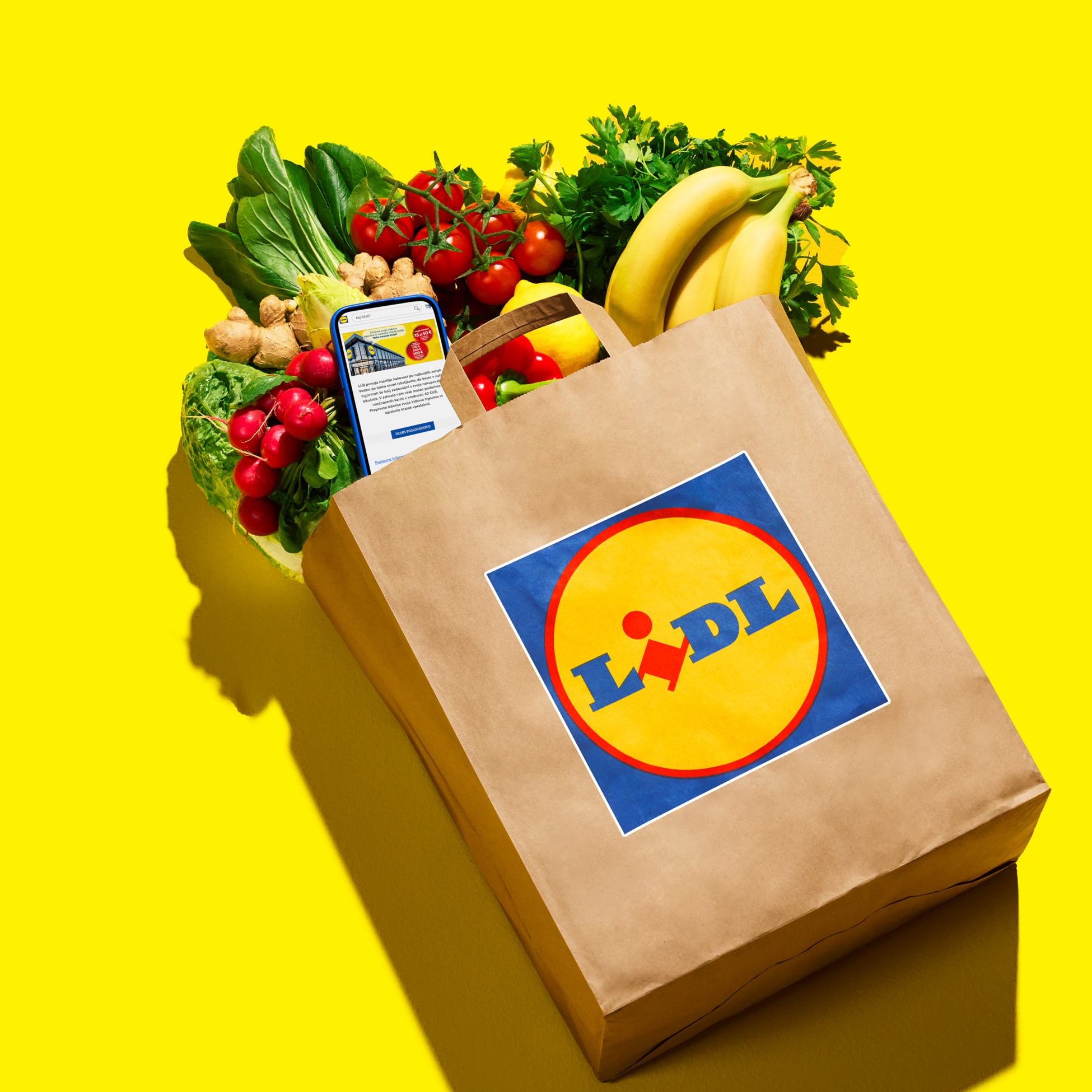 Papirnata vrećica s logotipom trgovine, puna svježeg povrća i voća, s telefonom koji prikazuje aplikaciju za kupovinu.