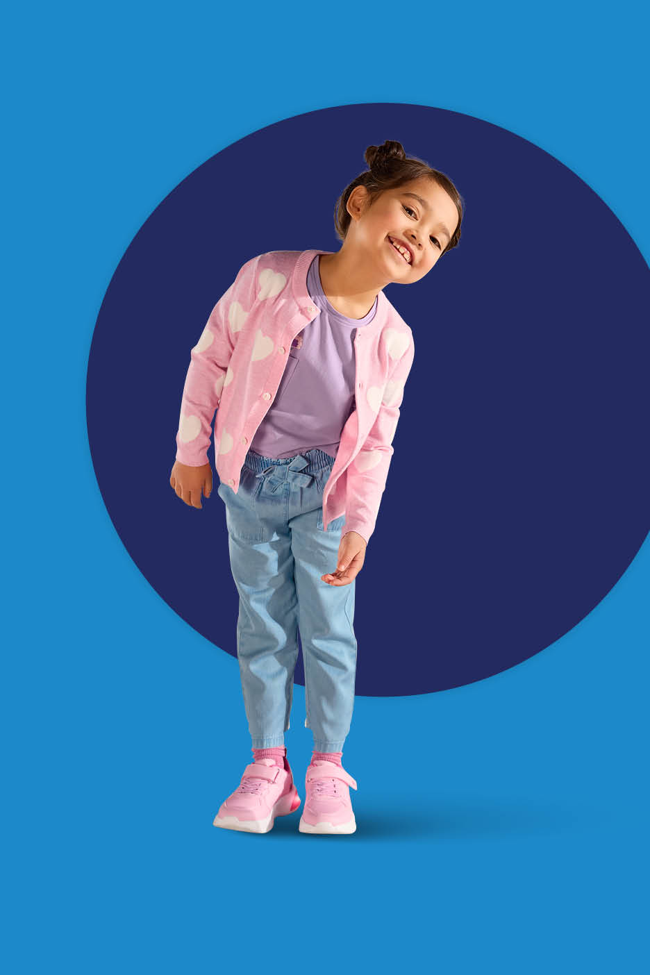 Djevojčica u ružičastom kardiganu sa srcima, ljubičastoj majici, trapericama i ružičastim tenisicama.