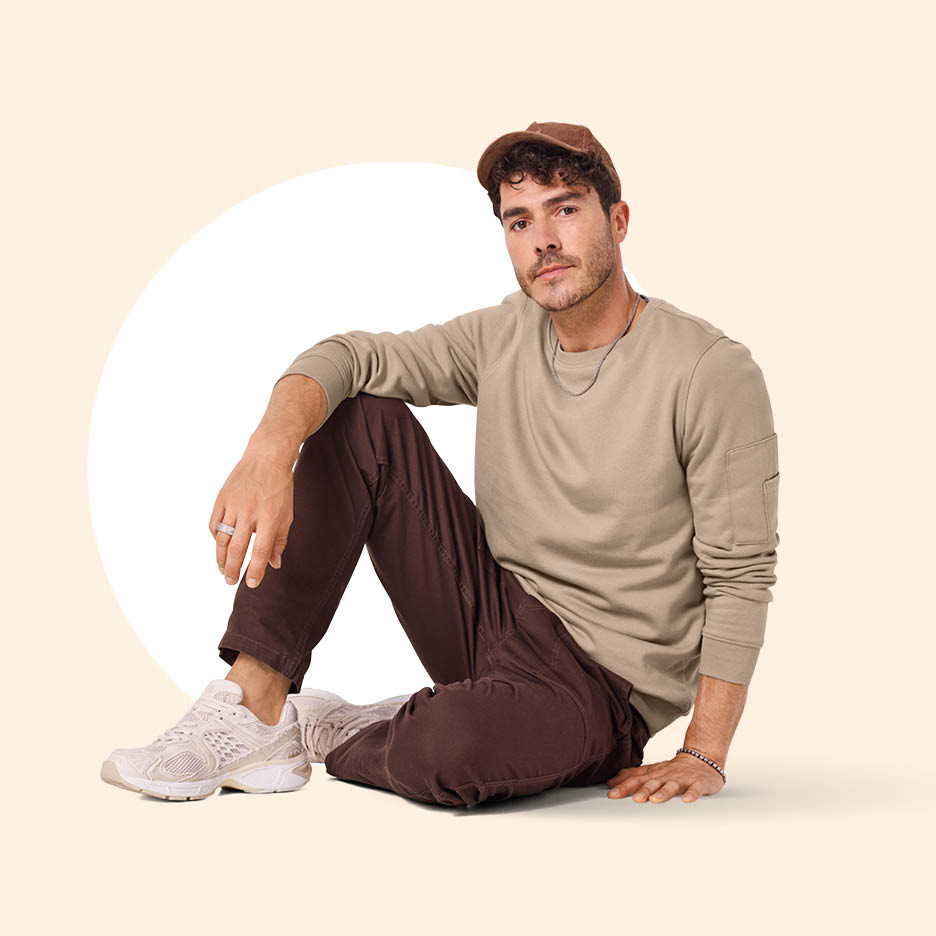 Muškarac u bež majici dugih rukava, smeđim hlačama i bež tenisicama sjedi na podu.