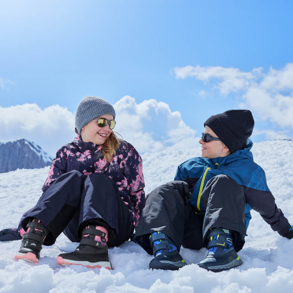 Dvoje djece u skijaškim jaknama, hlačama i čizmama sjedi na snijegu i smije se.