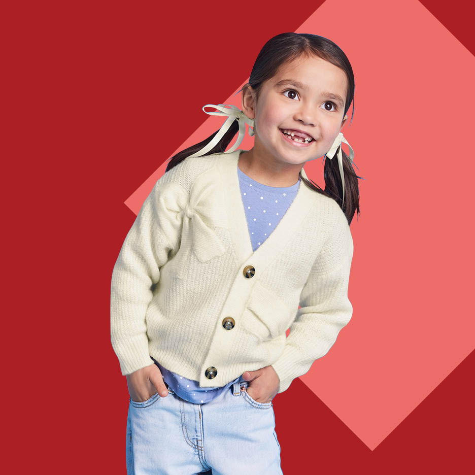 Djevojčica u krem kardiganu i plavoj majici s točkicama, s pletenicama i mašnama.