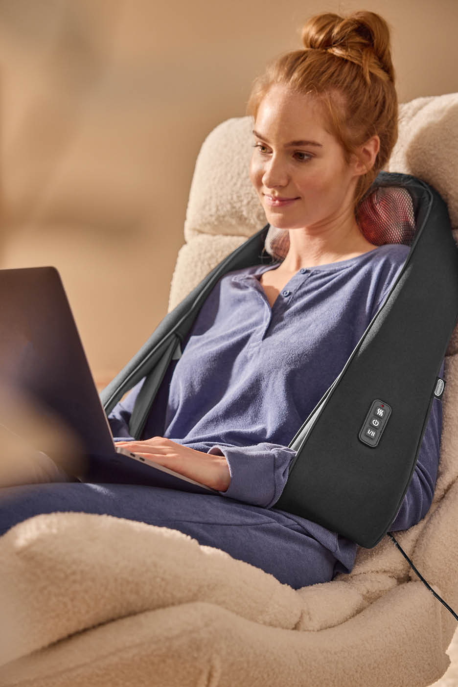 Žena s masažerom za vrat i ramena opušta se dok gleda u prijenosno računalo.