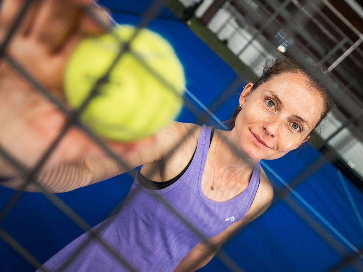 Žena u sportskoj odjeći drži padel lopticu kroz mrežu, plavi teren u pozadini.