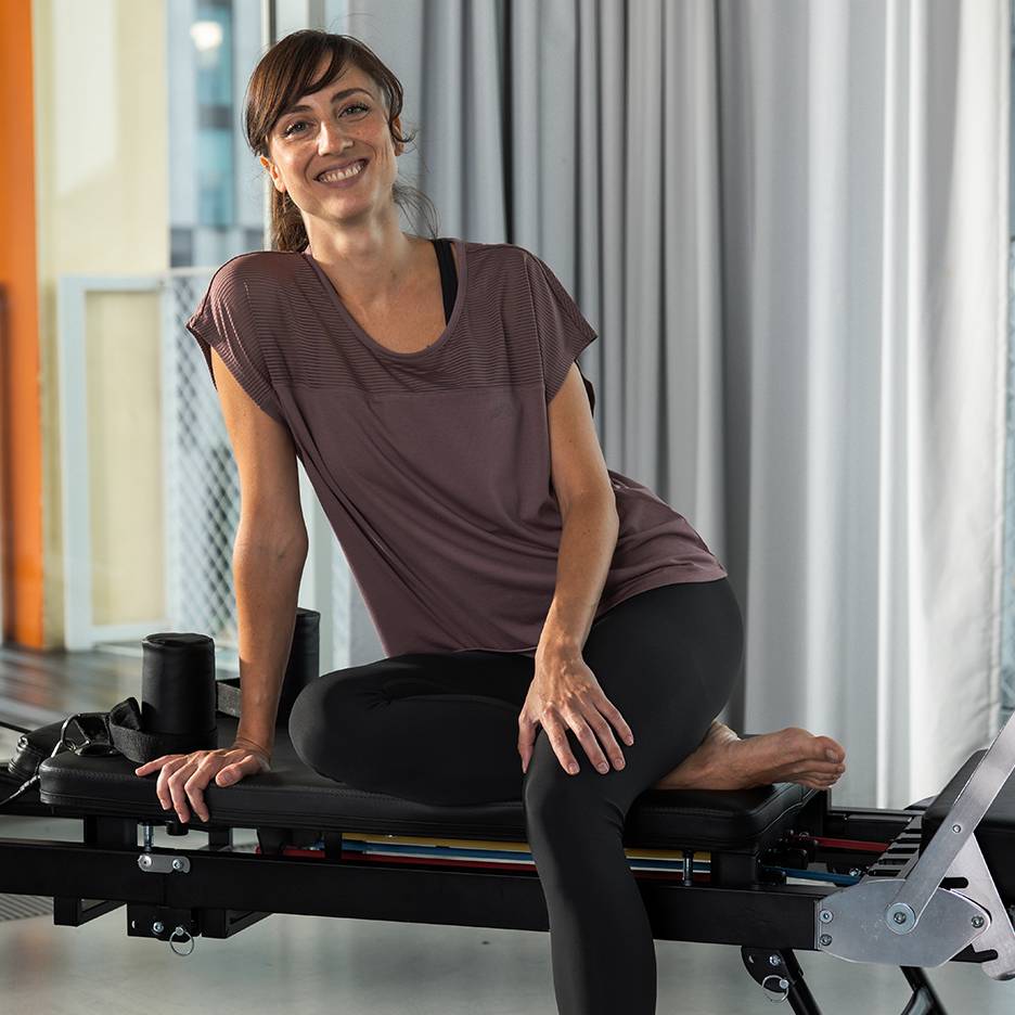 Žena u sportskoj odjeći nasmijana, sjedi na spravi za pilates.