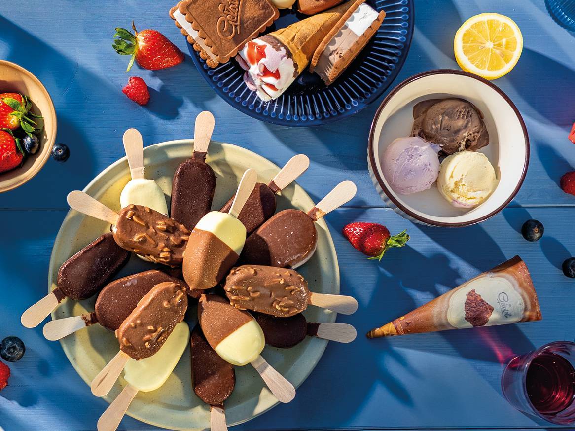 Raznovrsni sladoledi na štapiću, korneti i kuglice sladoleda na plavom stolu s voćem.