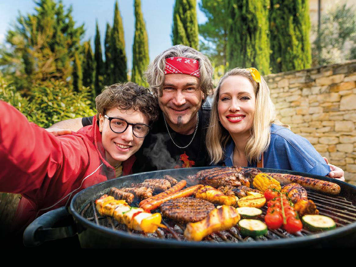 Obitelj se smije dok roštilja meso, kobasice i povrće na otvorenom.