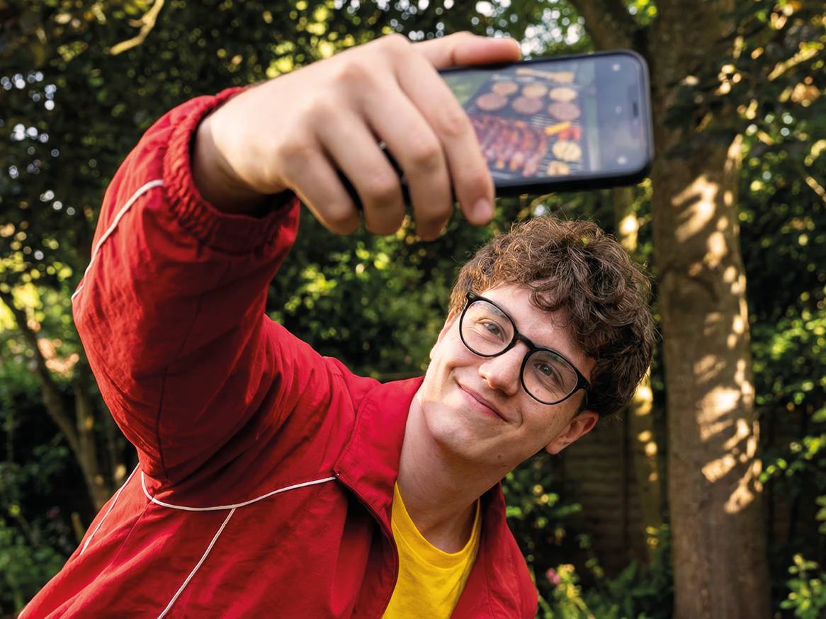 Mladić snima selfie s roštiljem punim mesa i povrća u pozadini.