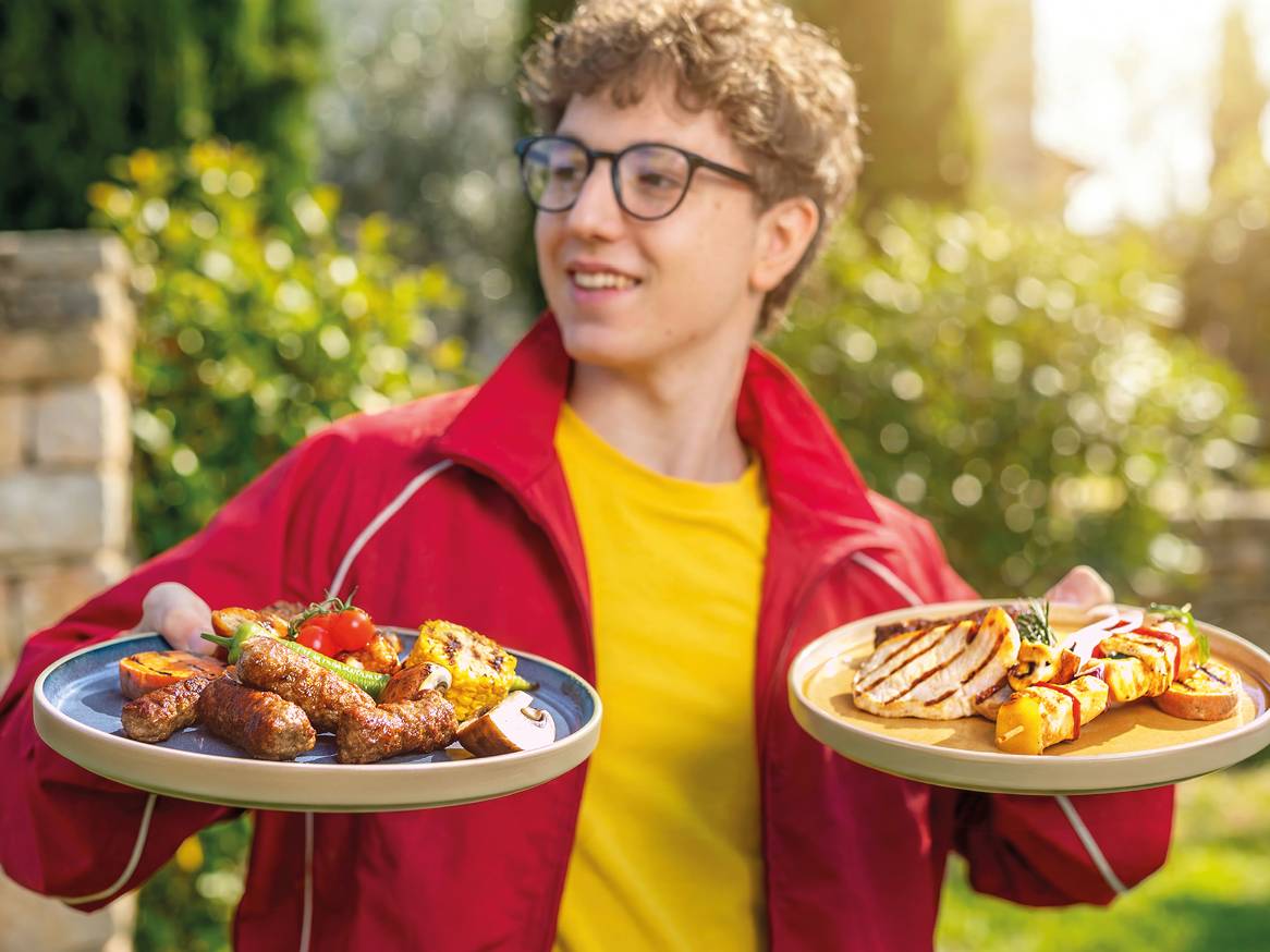 Mladić drži dva tanjura s roštiljem, uključujući ćevape, piletinu i povrće.