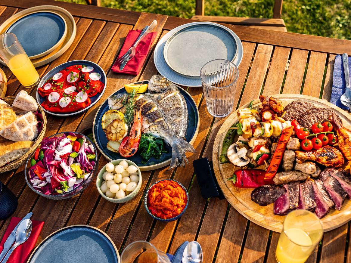 Roštilj s ribom, mesom, salatama i pićima na drvenom stolu na otvorenom.